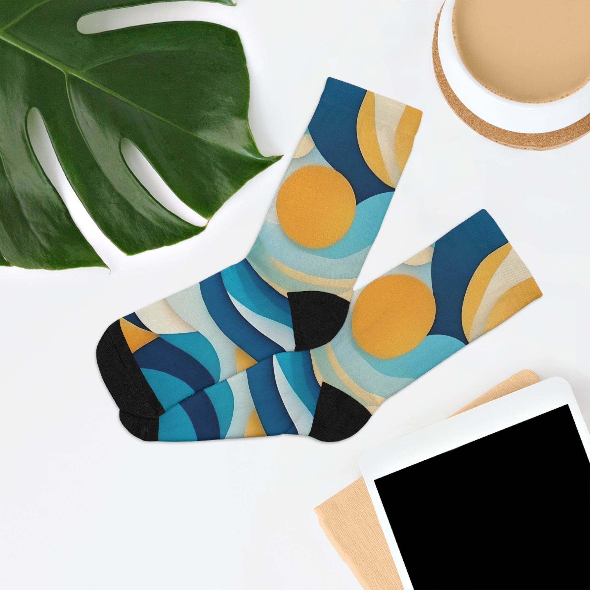 Printify eco-friendly recycled socks with colorful blue and yellow abstract pattern laid flat with tablet and plant