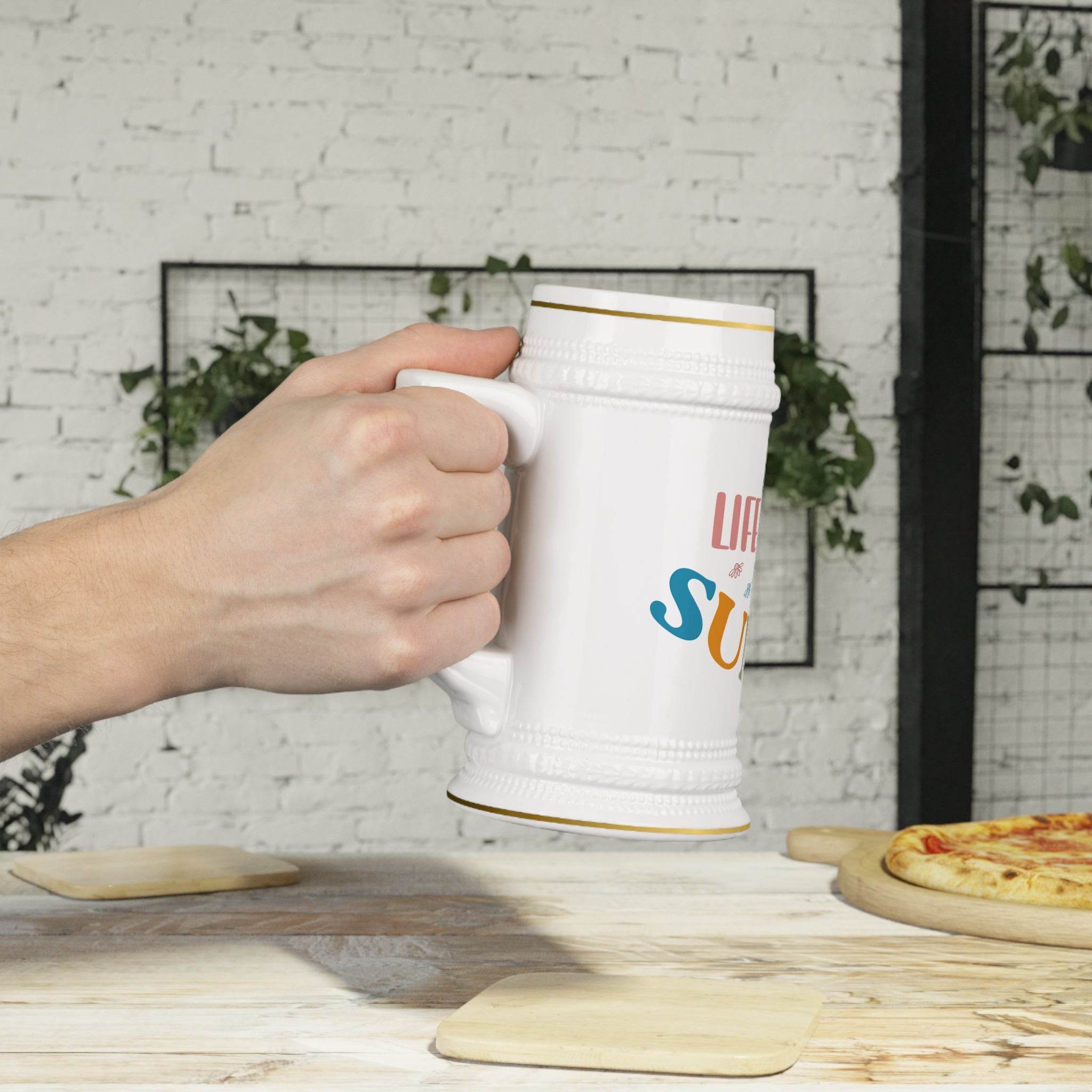 Hand holding a white eco-friendly beer stein mug by Printify with colorful text on a wooden table