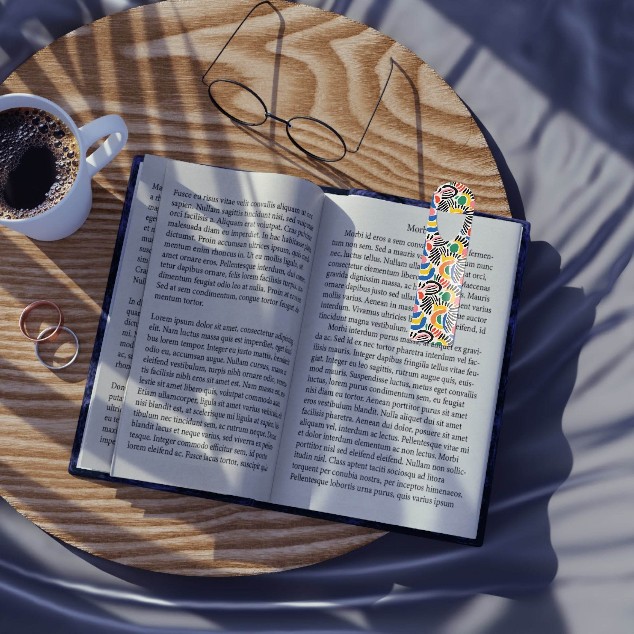 Open book with eco-friendly art bookmark featuring colorful abstract design on wooden table with glasses and coffee cup