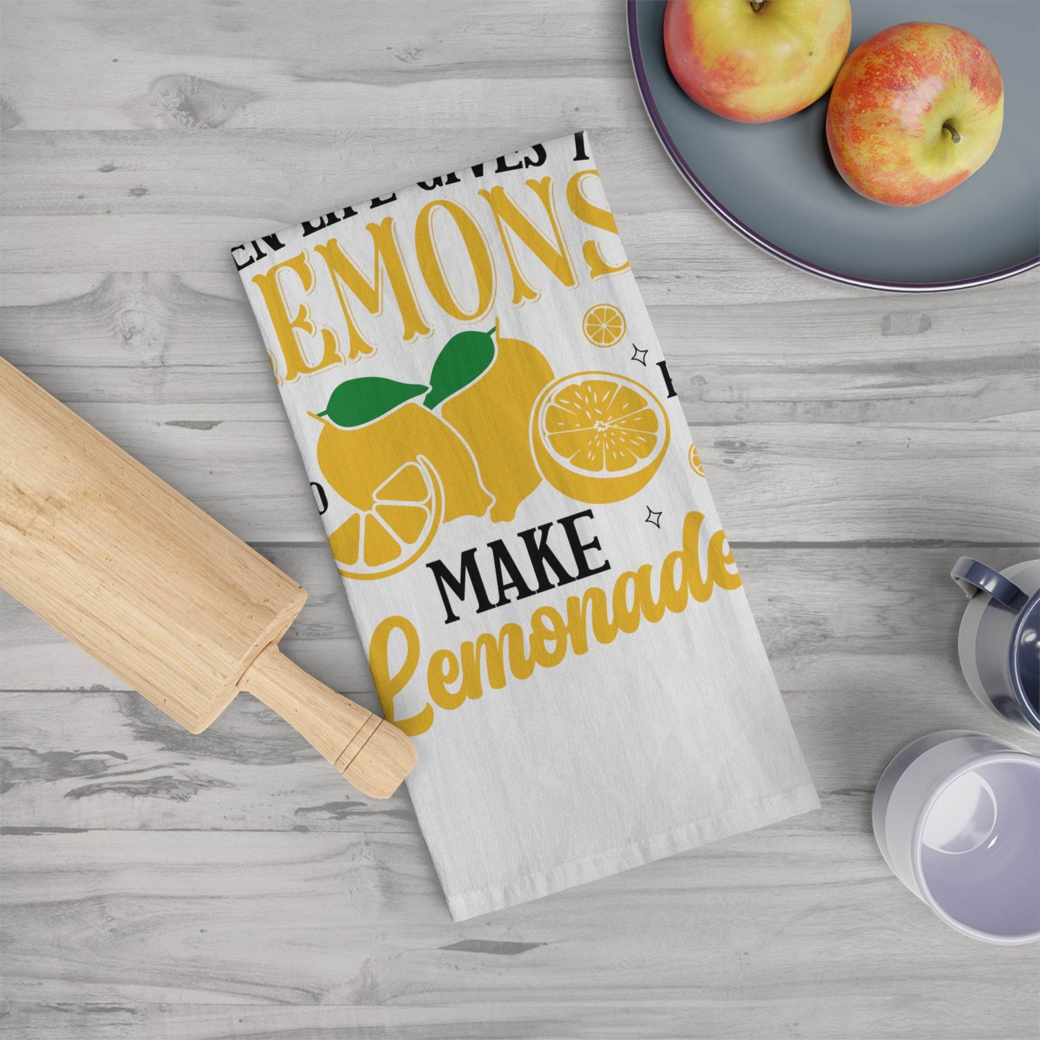 Lemonade themed tea towel with yellow lemons and text on white fabric next to a rolling pin and apples