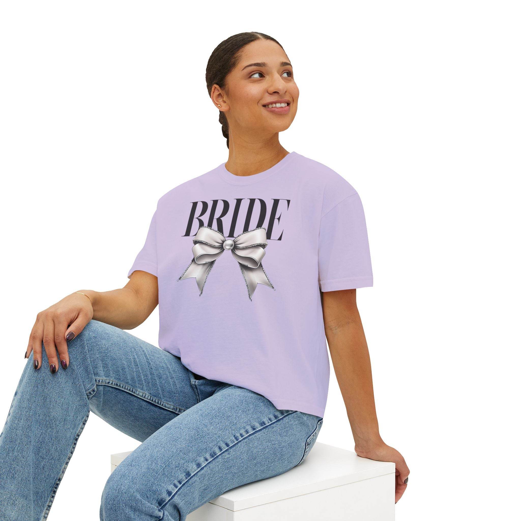 Woman wearing a Printify eco-friendly bridal t-shirt in lavender with BRIDE text and silver bow seated in jeans