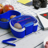 Blue reusable lunch container with clear lid and colorful text, holding fruits and nuts on a picnic table