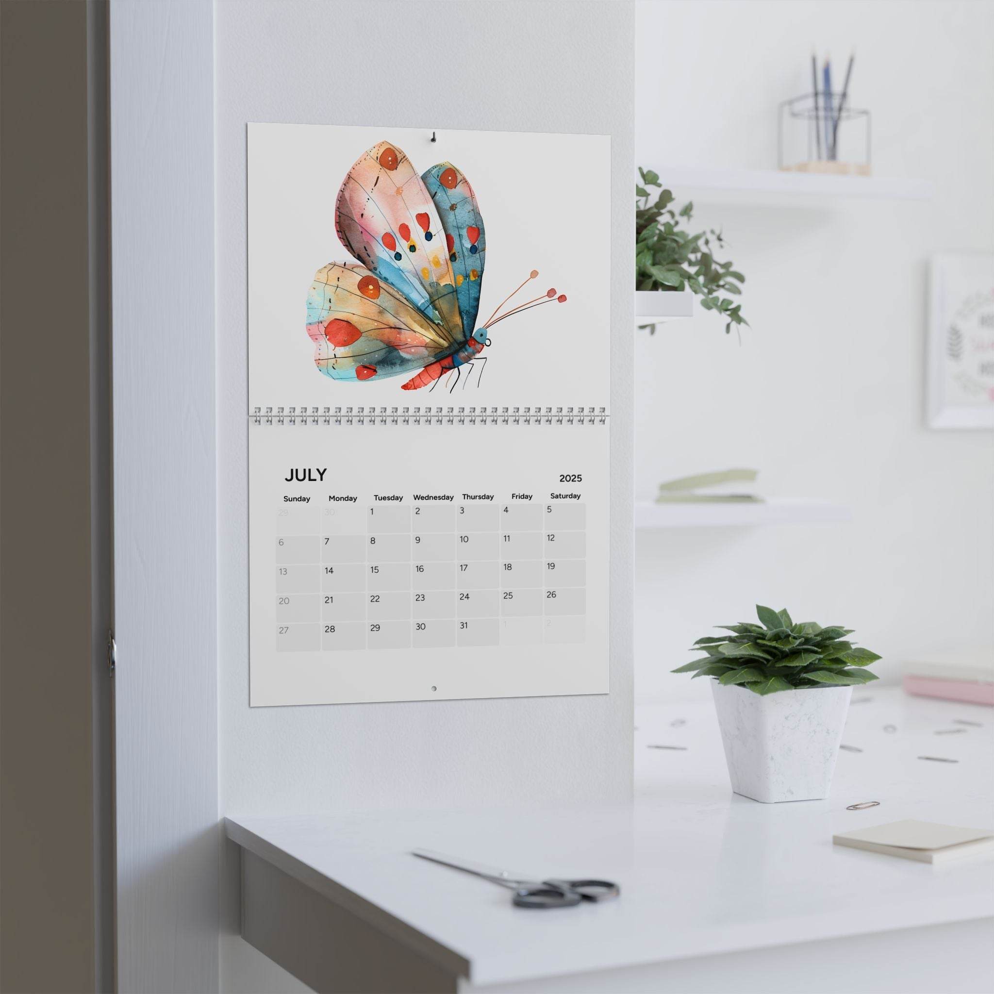 Printify butterfly design 2025 calendar hanging on wall above white desk with plant and scissors