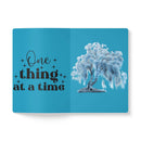 Open eco-friendly softcover journal with blue cover featuring one thing at a time quote and tree illustration
