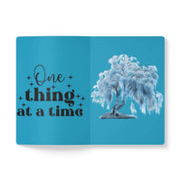Open eco-friendly softcover journal with blue cover featuring one thing at a time quote and tree illustration