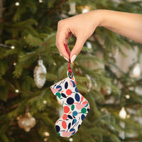Hand holding colorful eco-friendly wooden ornaments shaped like a Christmas stocking by a decorated tree