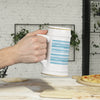 Hand holding a white ceramic stein mug with blue striped design and gold accents over wooden table