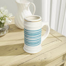 White ceramic stein mug with blue striped design on wooden table next to white pitcher and flowers